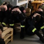 Firefighters in uniform with reflective stripes sit on benches, heads bowed in distress in a station setting.