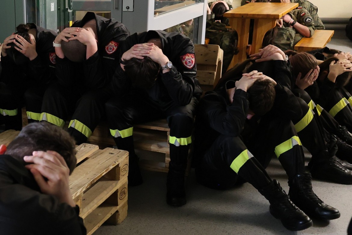 Firefighters in uniform with reflective stripes sit on benches, heads bowed in distress in a station setting.