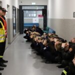 Two uniformed emergency responders stand at the end of a hallway as a group of students kneels against the wall with hands on their heads, participating in a drill or safety exercise.