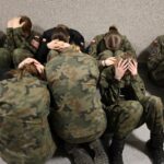 Group of soldiers in camouflage uniforms sitting against a wall, heads in hands, indicating stress or distress.