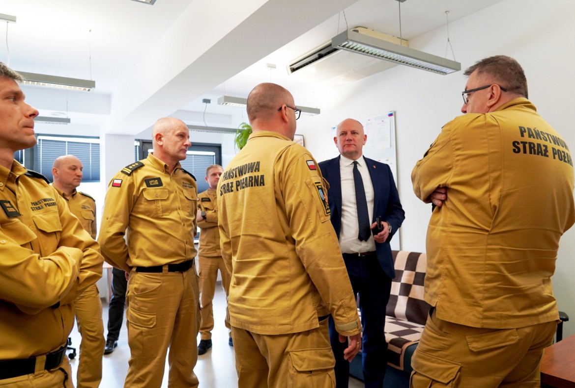 Group of Polish State Fire Service firefighters in tan uniforms listening to a man in a suit indoors.