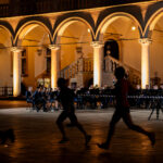 Evening outdoor orchestra under illuminated arches in a historic courtyard, players seated with music stands while blurred runners pass in the foreground.