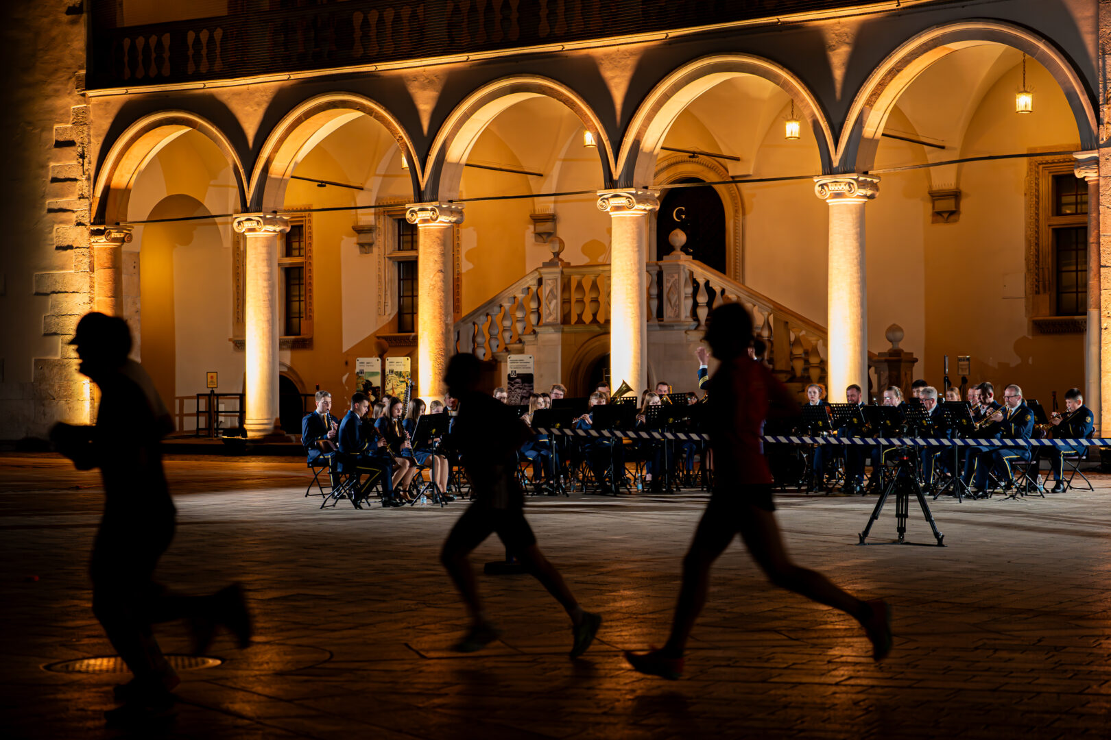 Evening outdoor orchestra under illuminated arches in a historic courtyard, players seated with music stands while blurred runners pass in the foreground.