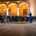 Blue-uniform wind band performing in a lit historic courtyard with arches; conductor directs from the front.