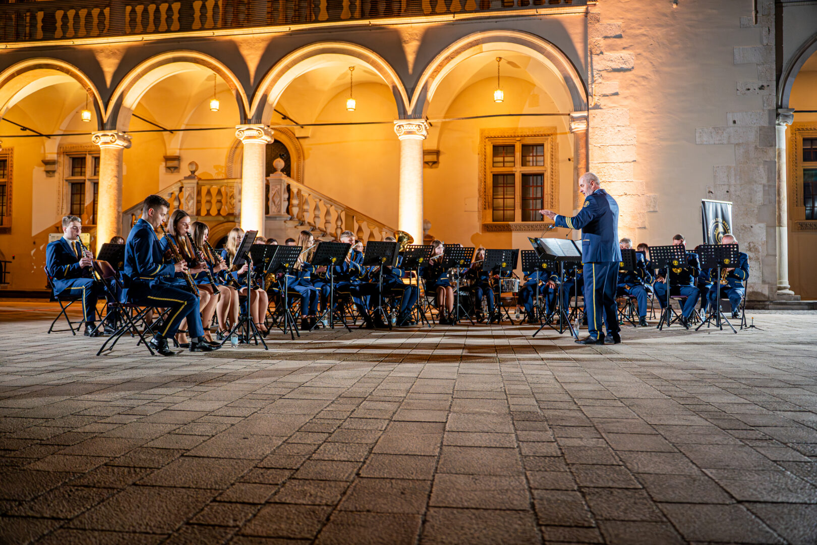 Blue-uniform wind band performing in a lit historic courtyard with arches; conductor directs from the front.