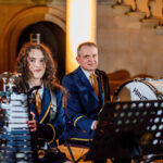 Two formally dressed percussionists sit with a xylophone (woman) and a large bass drum (man) during an outdoor performance at night.