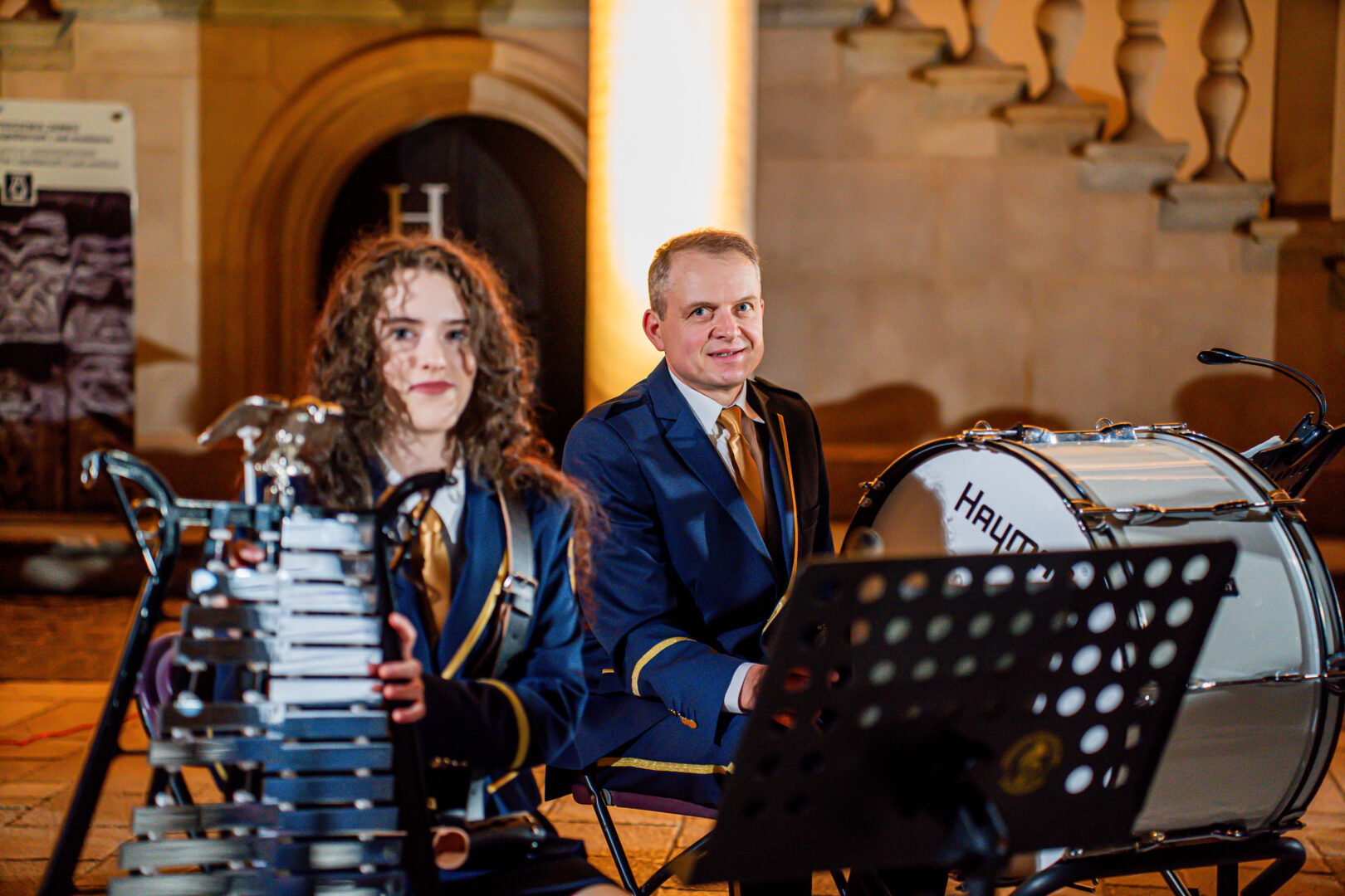 Two formally dressed percussionists sit with a xylophone (woman) and a large bass drum (man) during an outdoor performance at night.