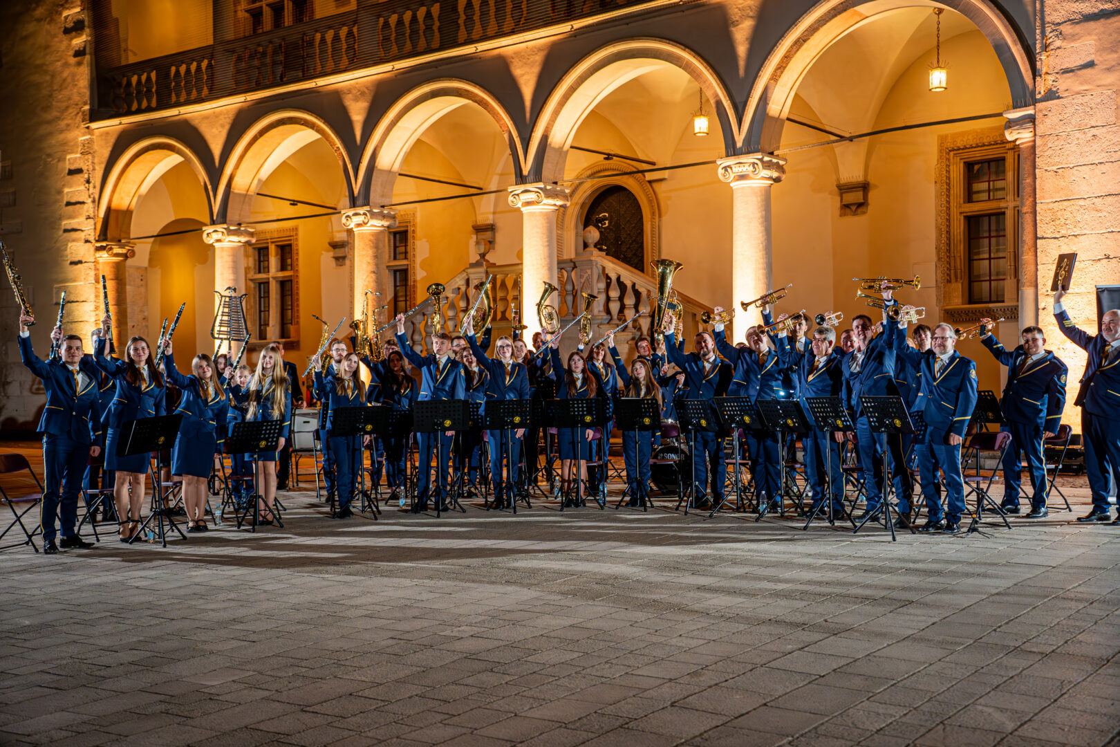 Brass band in blue uniforms raises instruments in a warmly lit arched courtyard at night.