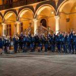 Large brass band in blue uniforms raises their instruments in a night-time outdoor performance under arched colonnades, courtyard setting.