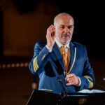 Man in a blue captain/'s uniform with gold stripes at a lectern, raising his right hand while speaking, with an open book nearby.