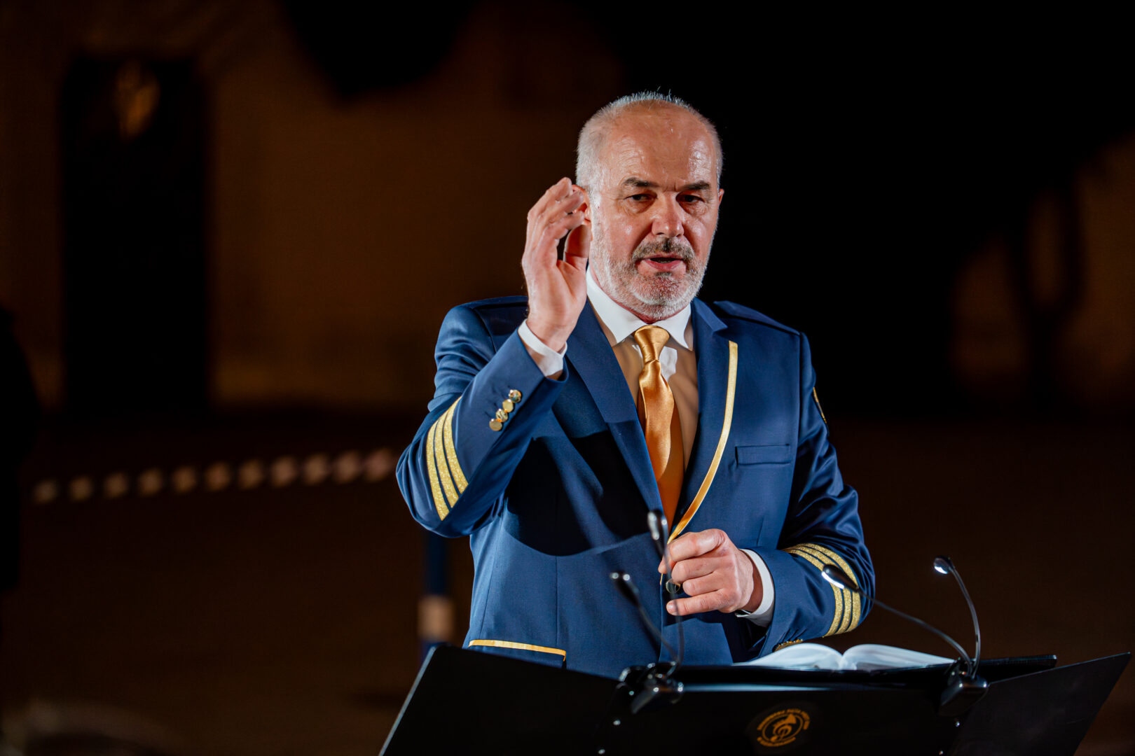 Man in a blue captain/'s uniform with gold stripes at a lectern, raising his right hand while speaking, with an open book nearby.
