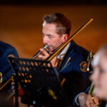 Male musician in a navy uniform playing a trombone, with a music stand in front and blurred bandmates in the background.