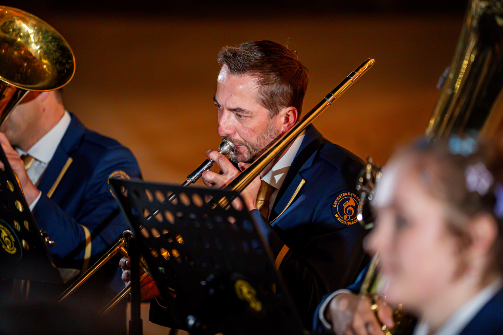 Male musician in a navy uniform playing a trombone, with a music stand in front and blurred bandmates in the background.