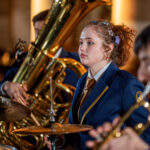 Young female drummer in a navy blazer and gold tie, focused on playing the drums in a band performance with brass instruments nearby.