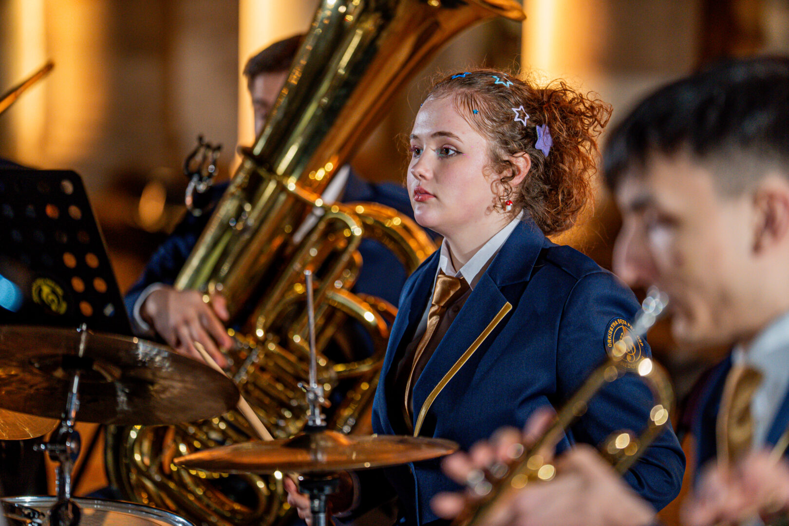 Young female drummer in a navy blazer and gold tie, focused on playing the drums in a band performance with brass instruments nearby.