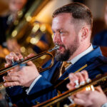 Close-up of a bearded trumpeter in a blue suit playing a trumpet during a band performance, with warm stage lights in the background.