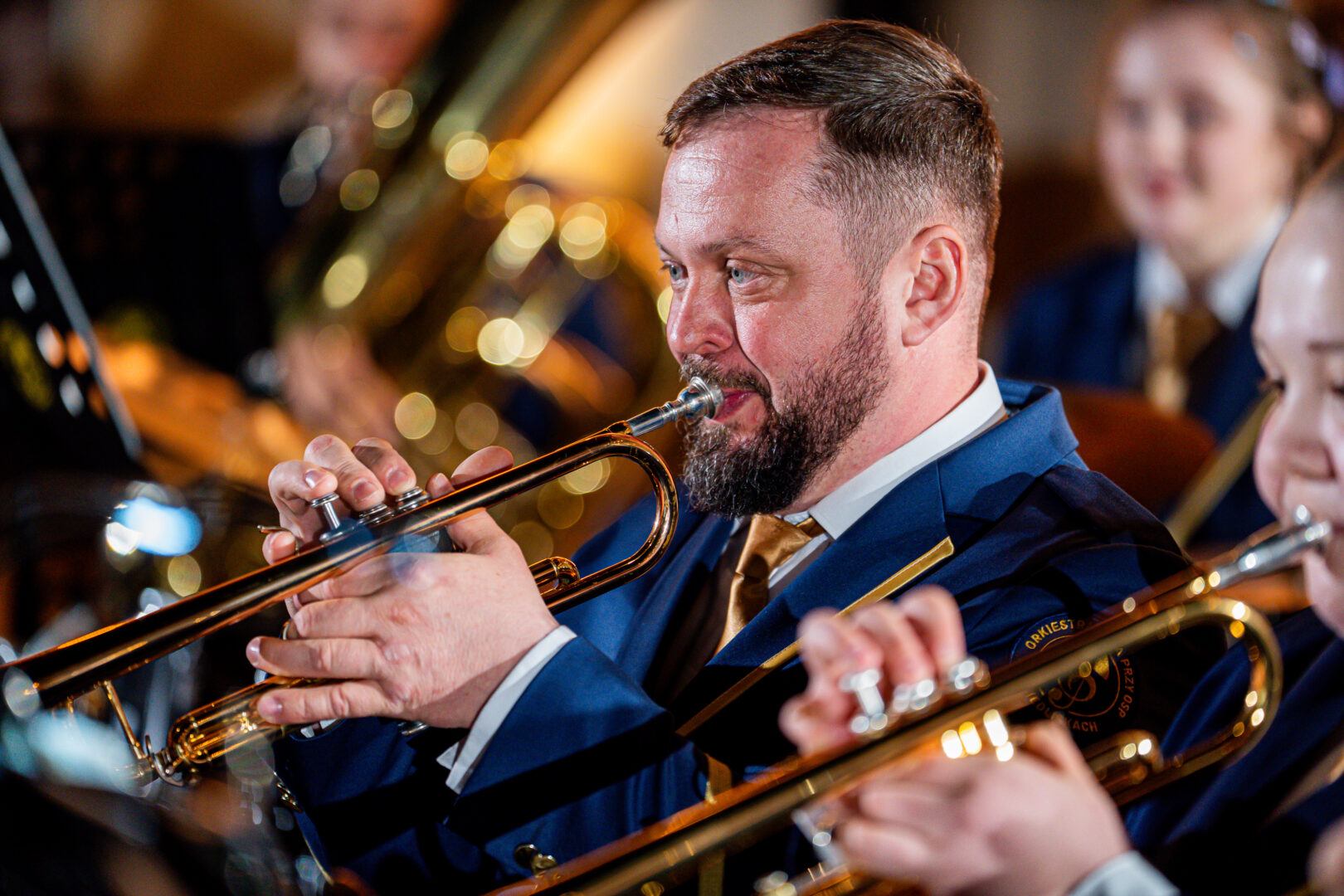 Close-up of a bearded trumpeter in a blue suit playing a trumpet during a band performance, with warm stage lights in the background.
