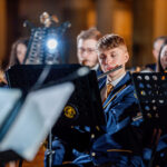 Young musicians in blue uniforms playing flutes, with music stands and stage lighting in the background.
