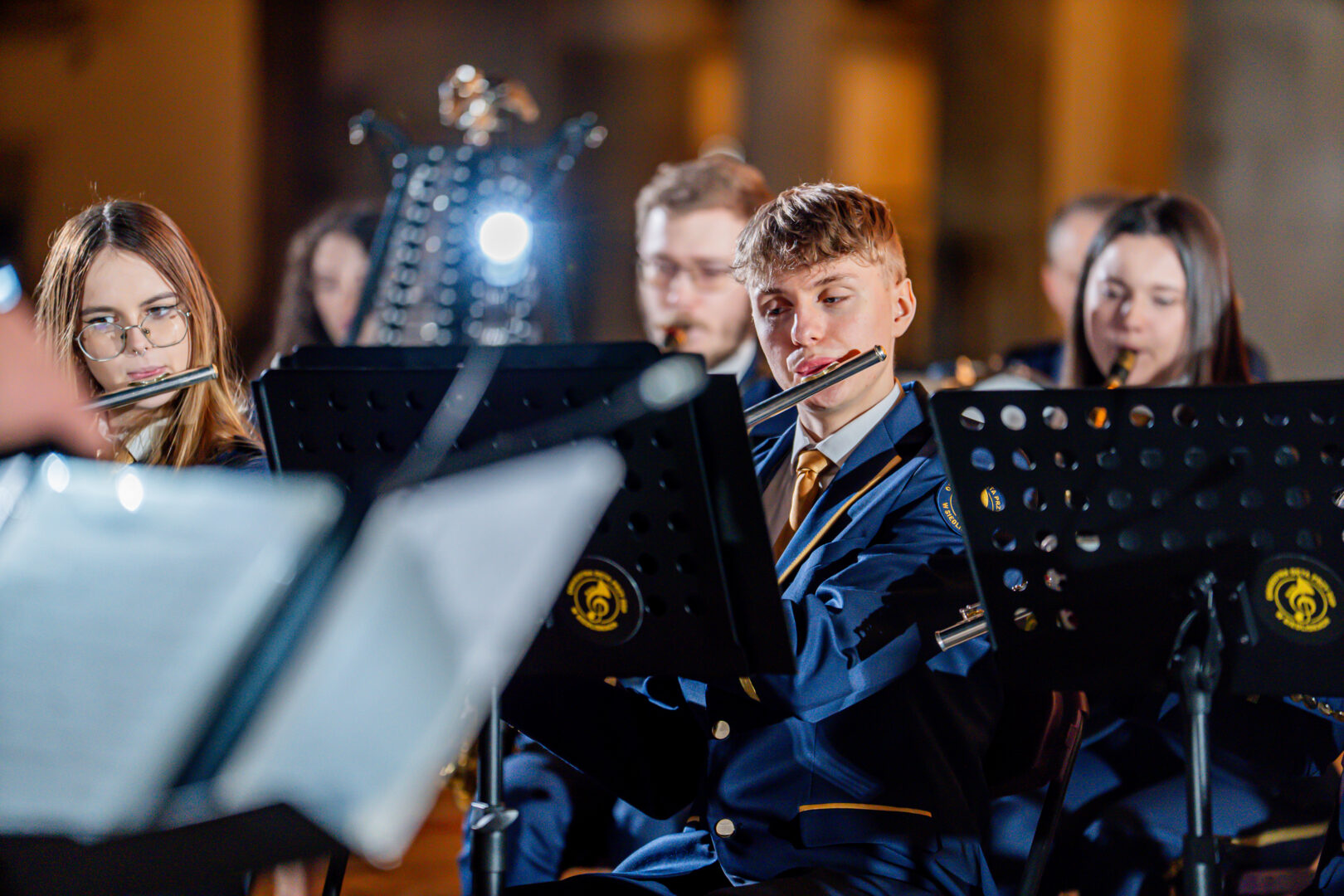 Young musicians in blue uniforms playing flutes, with music stands and stage lighting in the background.