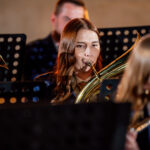 Female musician playing a brass instrument (tuba/euphonium) in a concert band, with music stands and other players visible behind her.