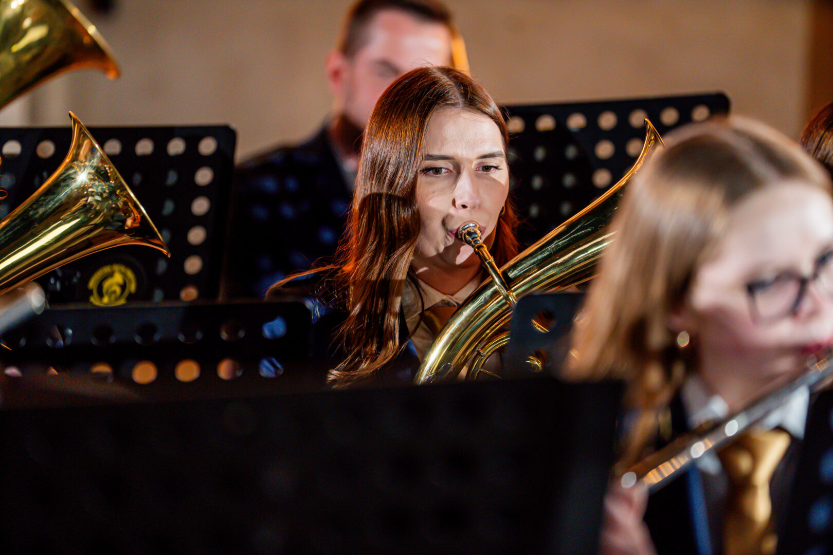 Female musician playing a brass instrument (tuba/euphonium) in a concert band, with music stands and other players visible behind her.
