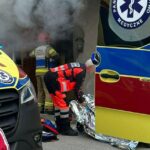 Emergency responders in reflective gear work outside a doorway as smoke rises; a medic unpacks a silver emergency blanket beside an ambulance.
