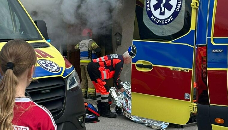 Emergency responders in reflective gear work outside a doorway as smoke rises; a medic unpacks a silver emergency blanket beside an ambulance.