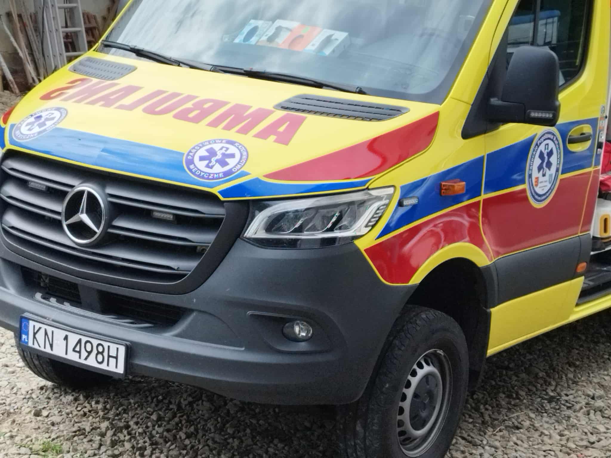 Front of a bright yellow ambulance with blue and red accents, large red 'AMBULANS' text, Star of Life sticker, and Polish license plate KN-1498H.
