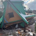 Collapsed wooden house with a green metal roof and scattered debris on the ground after a collapse, cloudy sky above.