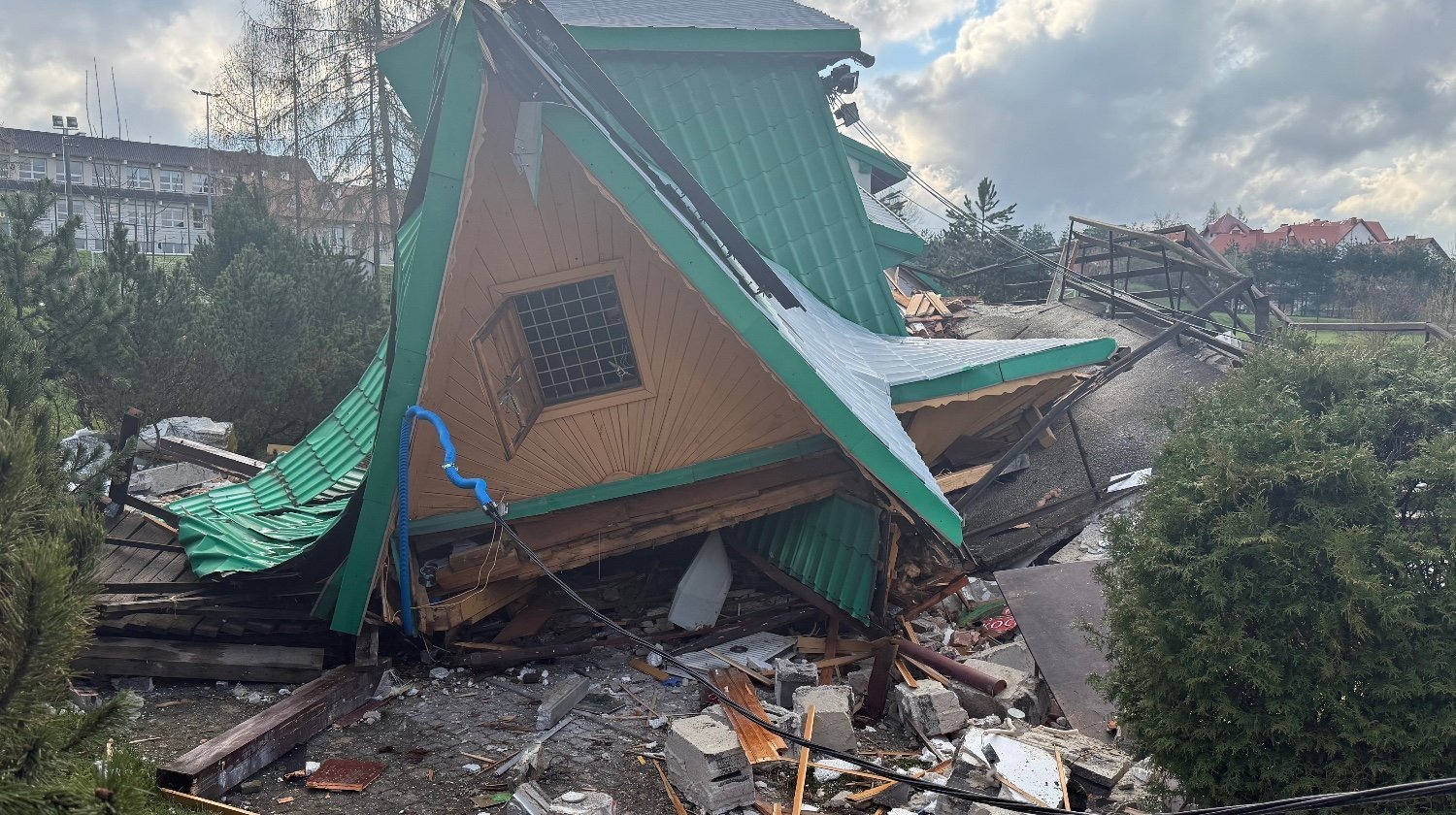 Collapsed wooden house with a green metal roof and scattered debris on the ground after a collapse, cloudy sky above.
