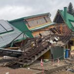 Partially collapsed house with a green metal roof, exposed brick and broken stairs amid rubble and caution tape outside.