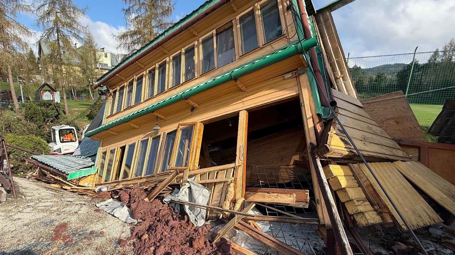 A partially collapsed wooden building tilted onto its side with scattered planks and debris around the structure, a small construction vehicle in the background on a grassy hill.