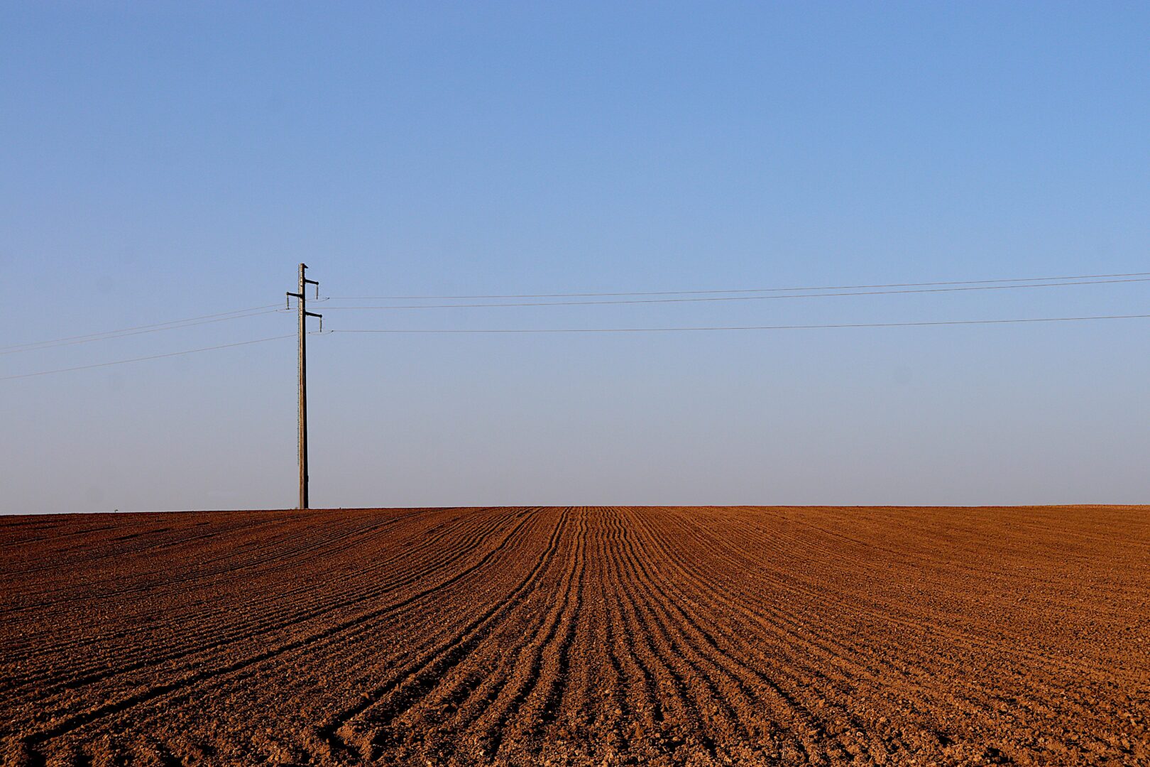 Single utility pole with power lines across a flat plowed field under a clear blue sky.