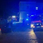 Night street with blue emergency lights from fire trucks and police cars; vehicles parked along a cobblestone road at night.