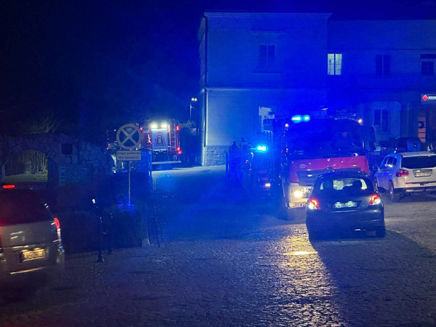 Night street with blue emergency lights from fire trucks and police cars; vehicles parked along a cobblestone road at night.