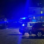 Nighttime street scene with blue emergency lights from police/fire vehicles and parked cars in the foreground.