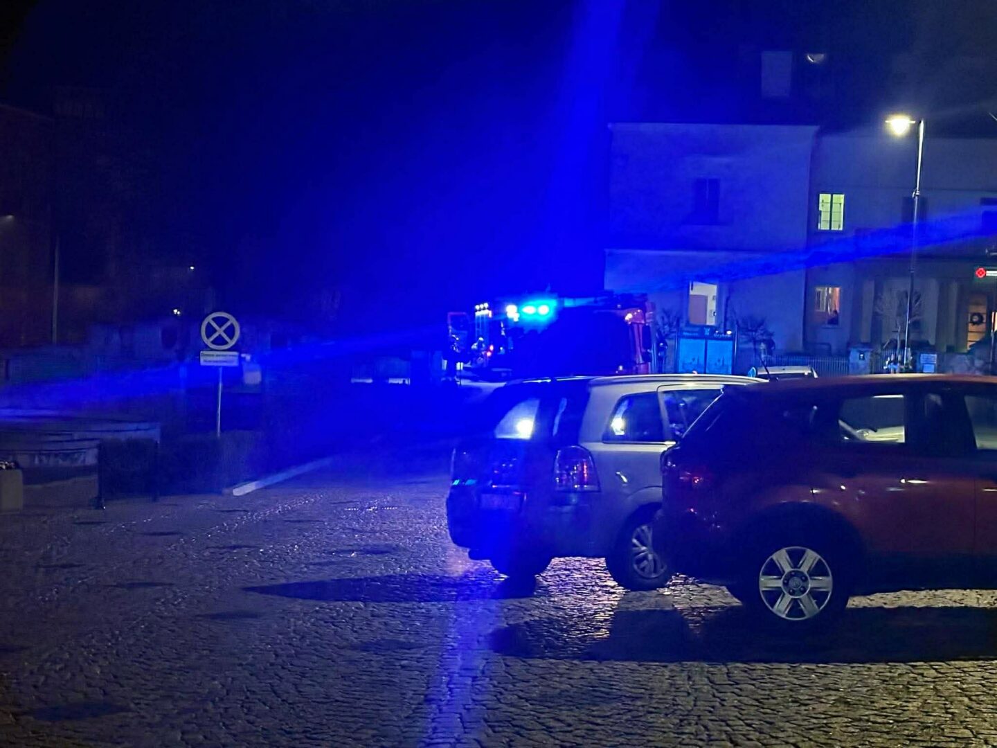 Nighttime street scene with blue emergency lights from police/fire vehicles and parked cars in the foreground.