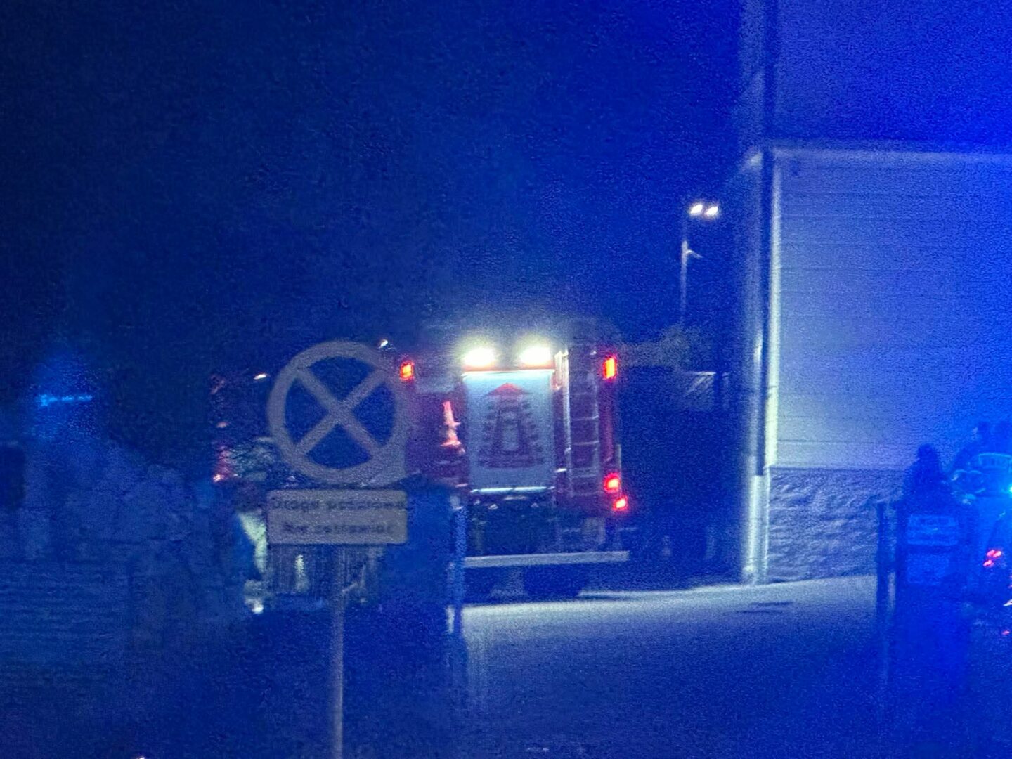 Night scene with a vehicle's rear lights on behind a gate and a large no-parking sign in the foreground, at dusk/nighttime UK-style street.