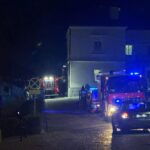 Nighttime street scene with blue emergency lights from a fire engine and an ambulance parked beside a building, several people gathered near the entrance.