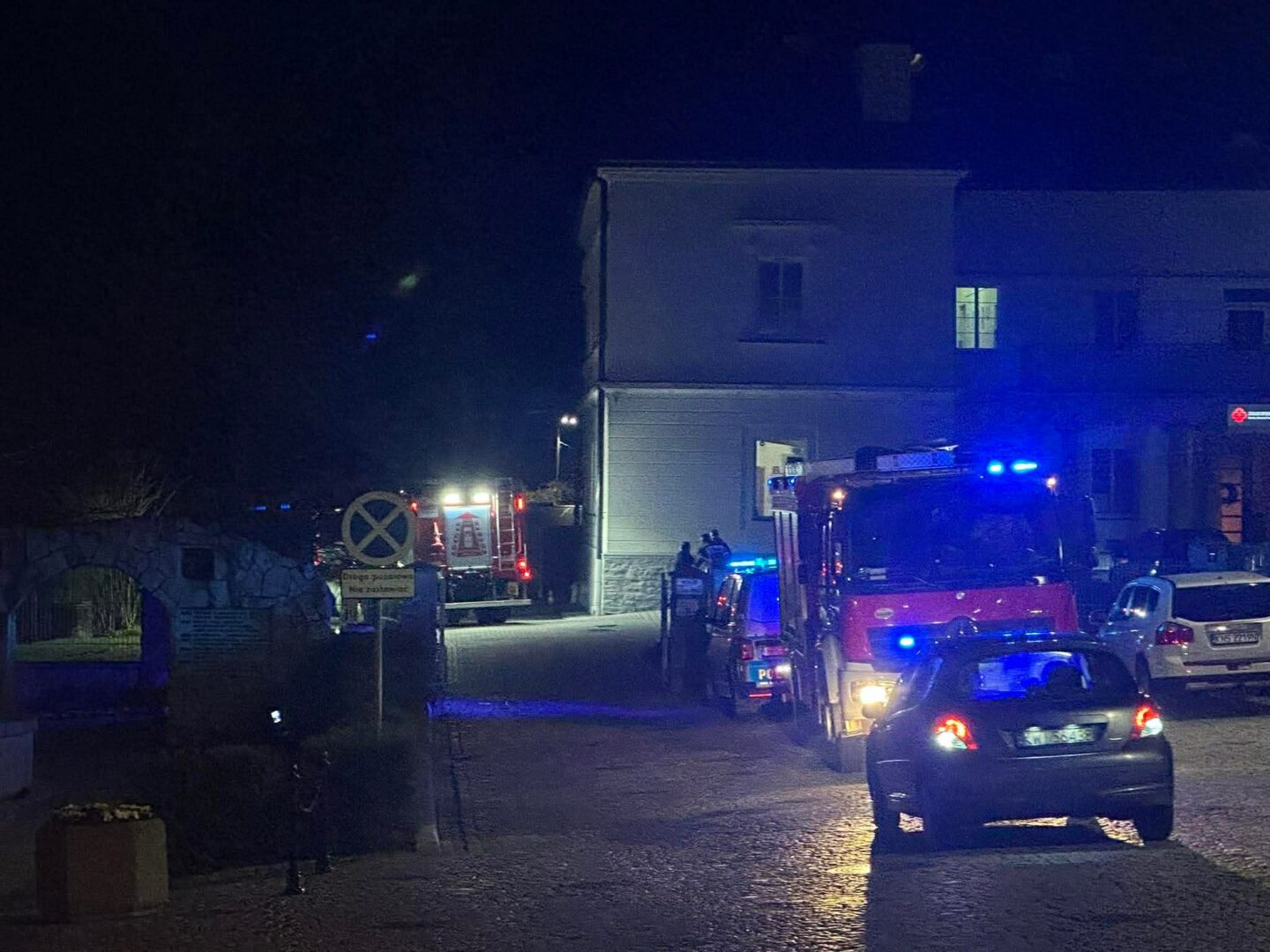 Nighttime street scene with blue emergency lights from a fire engine and an ambulance parked beside a building, several people gathered near the entrance.