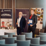 Two men in suits stand at a conference area with banners behind them; one holds a folder, the other a booklet; empty blue chairs in foreground.