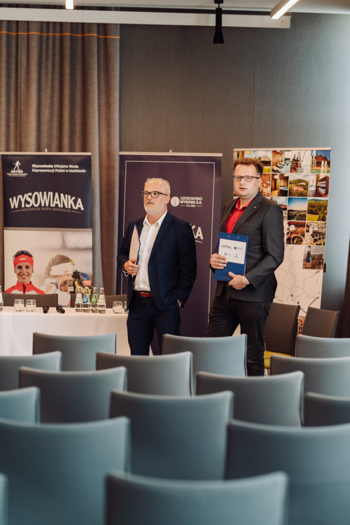 Two men in suits stand at a conference area with banners behind them; one holds a folder, the other a booklet; empty blue chairs in foreground.