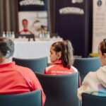 Audience view of a conference room with people seated; a young woman in a red jacket with white panels (UKS HANCZOWA) sits in the foreground facing a stage with banners and a panel table in the background.