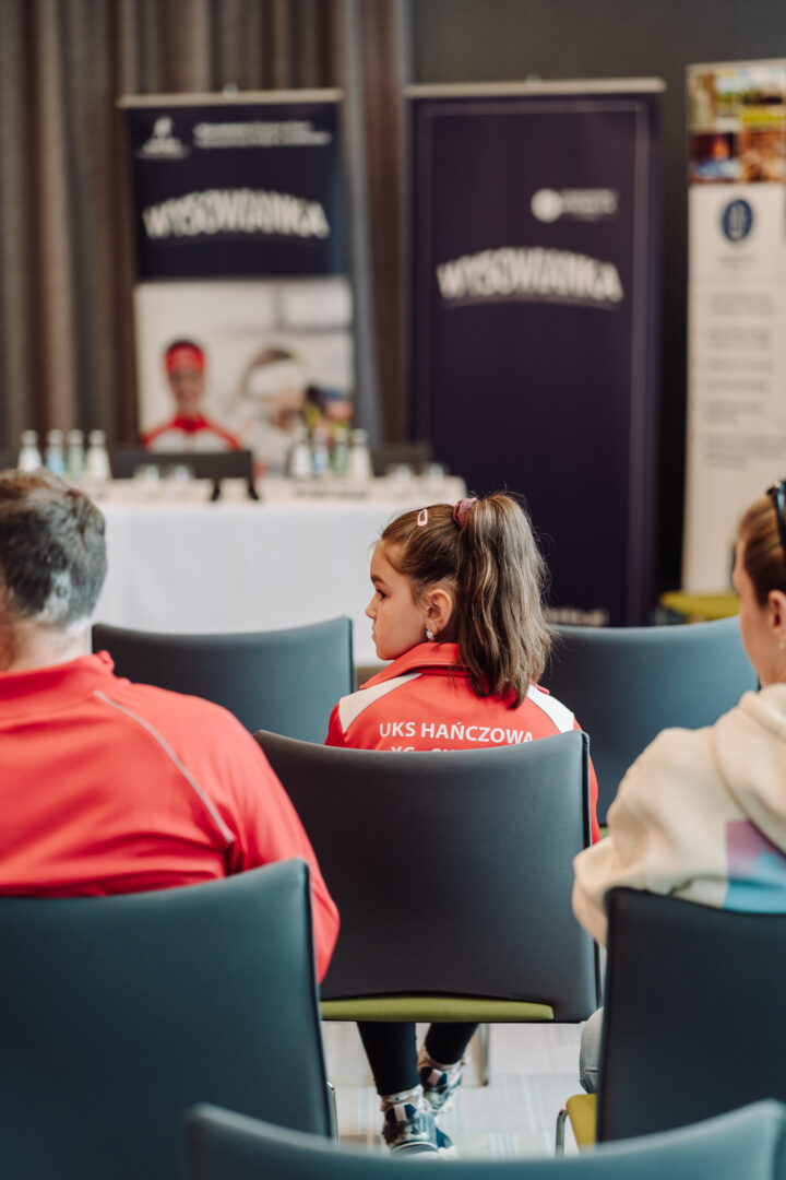 Audience view of a conference room with people seated; a young woman in a red jacket with white panels (UKS HANCZOWA) sits in the foreground facing a stage with banners and a panel table in the background.