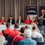 Panel of diverse speakers seated at a long table on a stage, with water bottles and microphones, audience in red shirts in front and banners behind them.