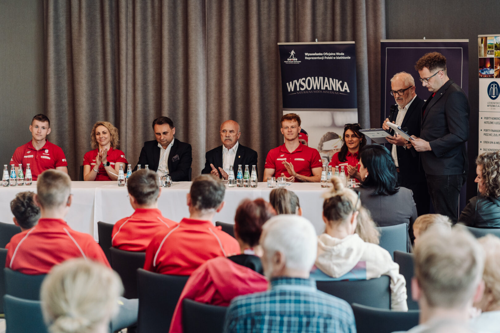 Panel of diverse speakers seated at a long table on a stage, with water bottles and microphones, audience in red shirts in front and banners behind them.