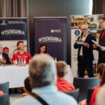 Panel discussion at a conference: seated panelists at a long table, two men standing with microphones addressing the audience, banners in the background.