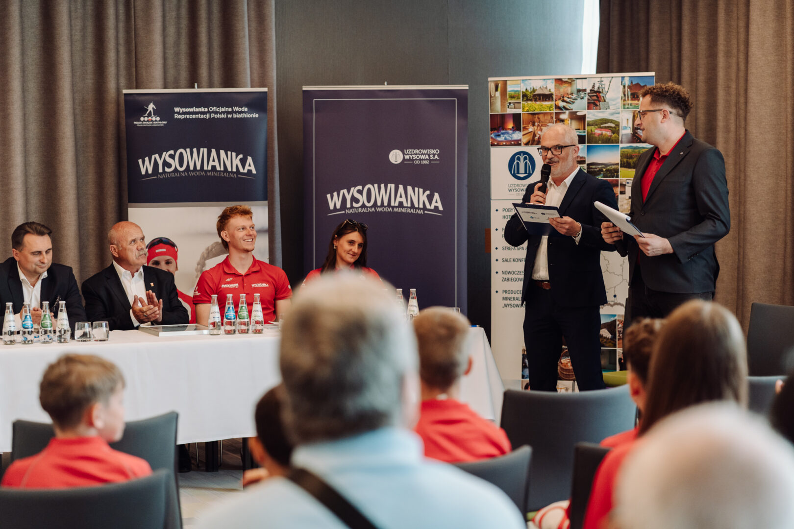Panel discussion at a conference: seated panelists at a long table, two men standing with microphones addressing the audience, banners in the background.