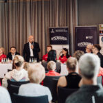 Man in a suit speaks into a microphone at a panel discussion, with banners and seated panelists behind him and an attentive audience in the foreground.