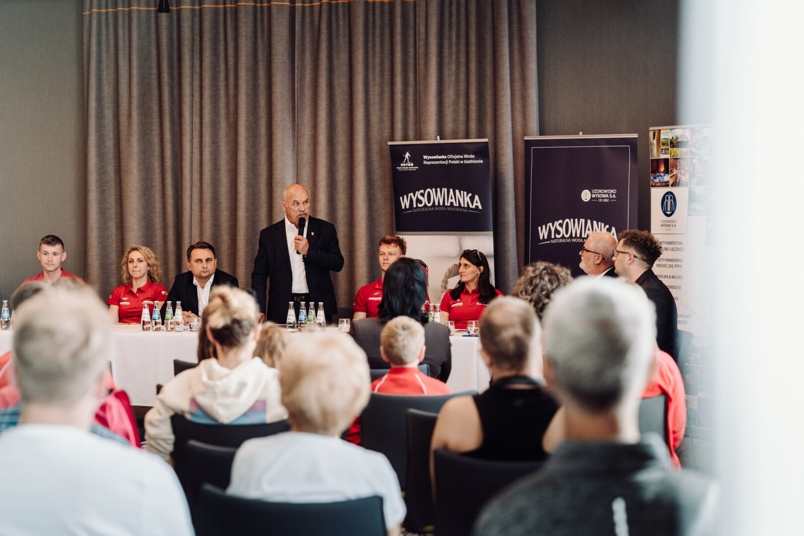 Man in a suit speaks into a microphone at a panel discussion, with banners and seated panelists behind him and an attentive audience in the foreground.